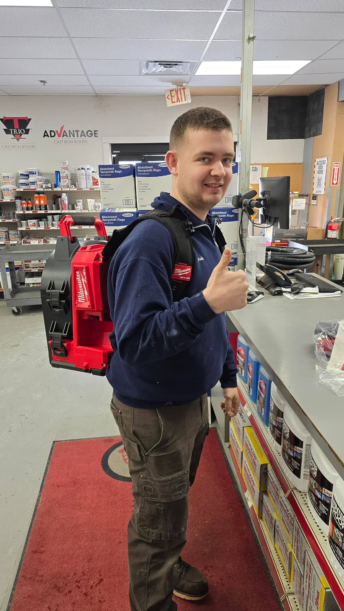 Man in a navy shirt wearing a red Milwaukee tool backpack, giving a thumbs up in a hardware store aisle.
